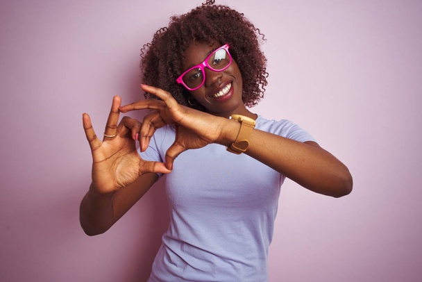 Młoda afrykańska afro kobieta w okularach na odizolowanym różowym tle, uśmiechnięta miłością, z symbolem serca i kształtem dłoni. Romantyczna koncepcja. - Zdjęcie, obraz