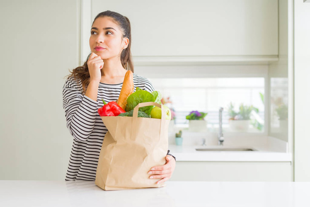 Fiatal nő gazdaság papírzacskót tele élelmiszereket komoly arca gondolkodás kérdés, nagyon zavaros ötlet - Fotó, kép
