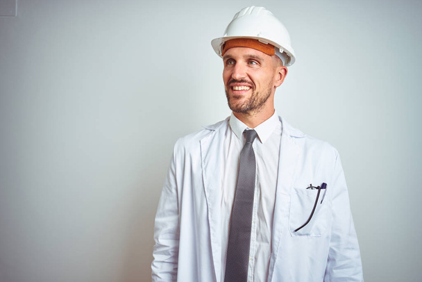 Young handsome engineer man wearing safety helmet over isolated background looking away to side with smile on face, natural expression. Laughing confident. - Foto, Imagem