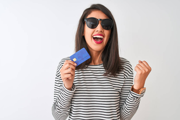 Young chinese woman wearing sunglasses holding credit card over isolated white background screaming proud and celebrating victory and success very excited, cheering emotion - Photo, image