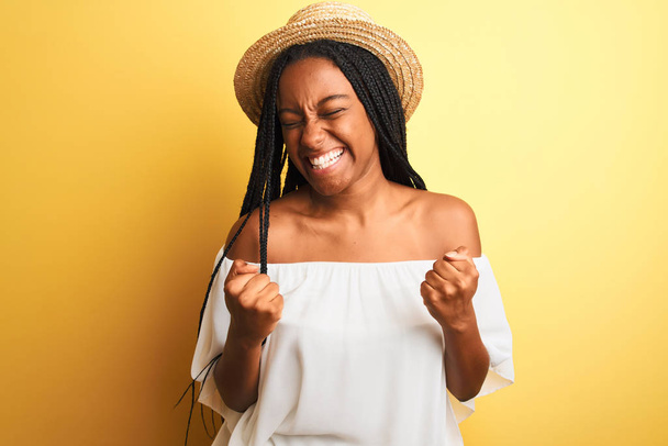 Mladá afro-americká žena v bílém tričku a klobouku na izolovaném žlutém pozadí velmi šťastná a nadšená dělá vítězný gesto se zdviženýma rukama, usmívá se a křičí po úspěchu. Koncept oslavy. - Fotografie, Obrázek