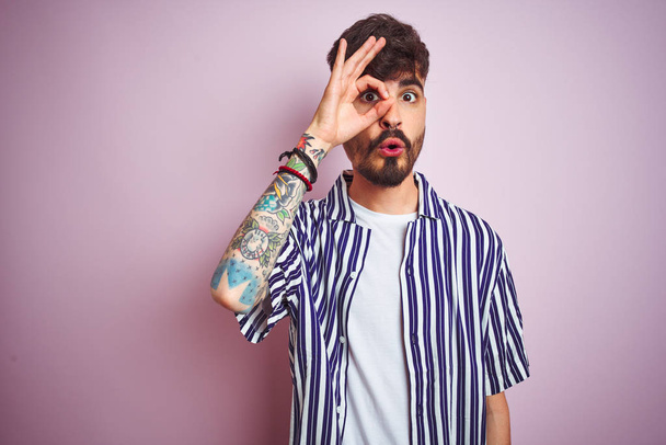 Jongeman met tatoeage met gestreept shirt over geïsoleerde roze achtergrond staan doen ok gebaar geschokt met verrast gezicht, oog kijken door vingers. Ongelovige uitdrukking. - Foto, afbeelding