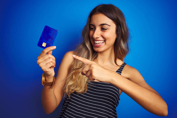 Молода красива жінка тримає кредитну картку над синім ізольованим тлом дуже щаслива, вказуючи рукою і пальцем
 - Фото, зображення