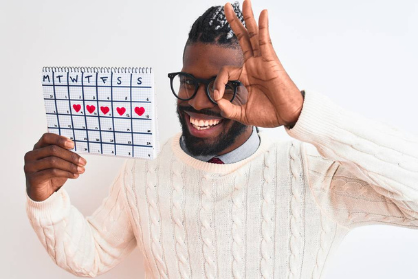 Homme afro-américain avec des tresses tenant calendrier période sur fond blanc isolé avec le visage heureux souriant faire ok signe avec la main sur l'oeil regardant à travers les doigts
 - Photo, image