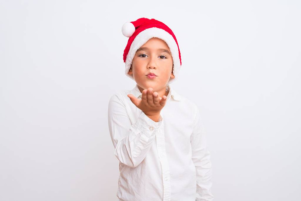 Hermoso niño con sombrero de Navidad Santa de pie sobre fondo blanco aislado mirando a la cámara soplando un beso con la mano en el aire siendo encantador y sexy. Expresión de amor
. - Foto, Imagen