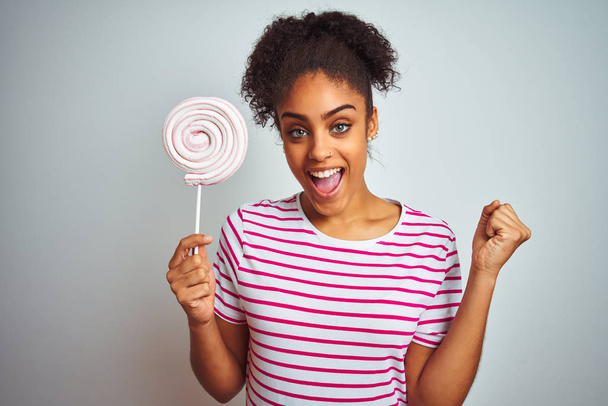 afroamericano adolescente donna mangiare caramelle colorate su sfondo bianco isolato urlando orgoglioso e celebrando la vittoria e il successo molto eccitato, emozione tifo
 - Foto, immagini