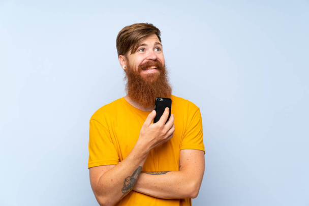 Рудий чоловік з довгою бородою з мобільним телефоном над ізольованою синьою стіною, дивлячись вгору, посміхаючись
 - Фото, зображення