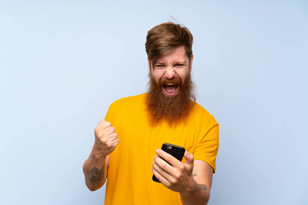Lucky Redhead man with long beard with a mobile over isolated blue wall - Photo, Image