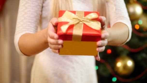 Closeup felvétel fiatal nő piros karácsonyi ajándékdoboz mellett álló díszített karácsonyfa a nappaliban. Családi adó és fogadó ajándékok a téli szünnapokon és ünnepségeken. - Felvétel, videó