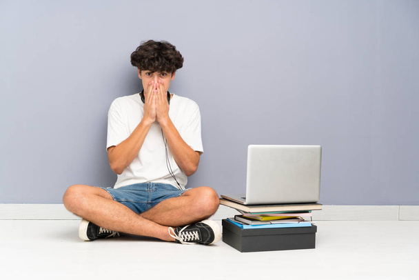 Fiatal férfi laptoppal ül a padlón, meglepett arckifejezéssel. - Fotó, kép