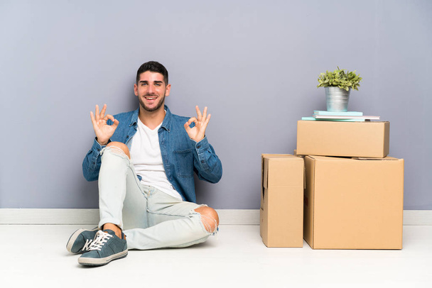 Handsome young man moving in new home among boxes showing an ok sign with fingers - Foto, Bild