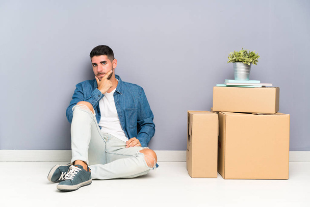 Красивый молодой человек переезжает в новый дом среди коробок, думая об идее
 - Фото, изображение