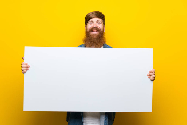 Roodharige man met lange baard over geïsoleerde gele muur met een leeg wit bordje voor het invoegen van een concept - Foto, afbeelding