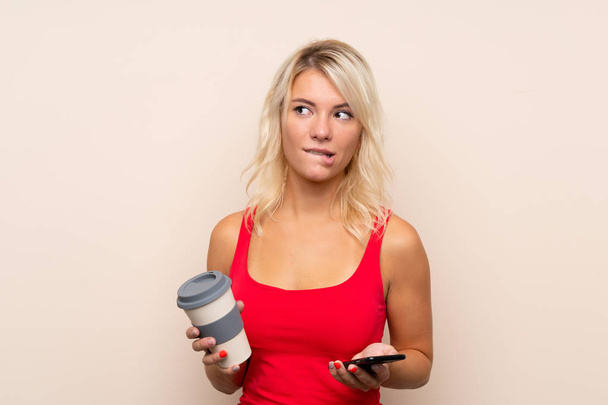 Young blonde woman over isolated background holding coffee to take away and a mobile while thinking something - Foto, Bild
