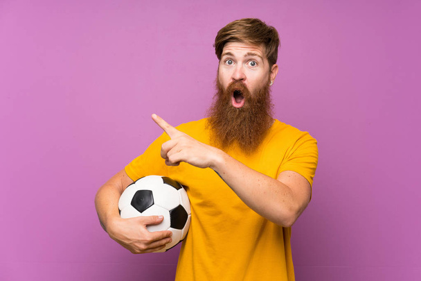 Vörös hajú férfi hosszú szakállal kezében egy focilabda felett elszigetelt lila fal meglepett és rámutatott oldalán - Fotó, kép