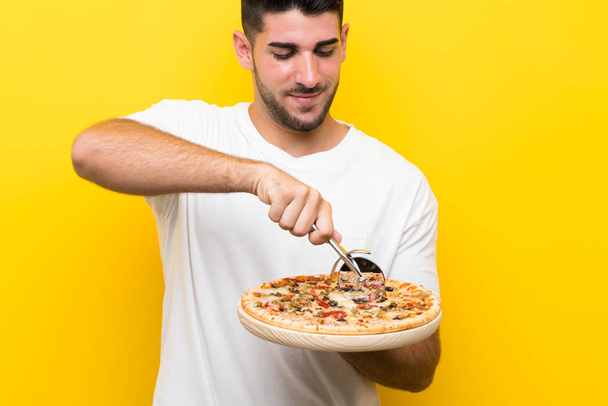 İzole sarı duvar üzerinde bir pizza tutan genç yakışıklı adam - Fotoğraf, Görsel