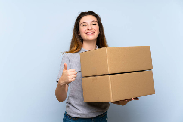 Young brunette girl over isolated blue background holding a box to move it to another site with thumb up - Photo, Image