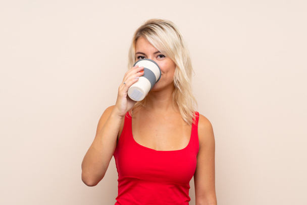 Young blonde woman over isolated background holding and drinking a coffee to take away - Foto, Bild