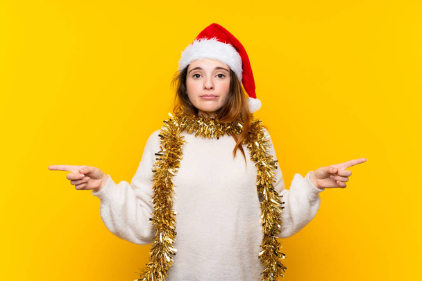 Girl with christmas hat over isolated yellow background pointing to the laterals having doubts - Photo, Image