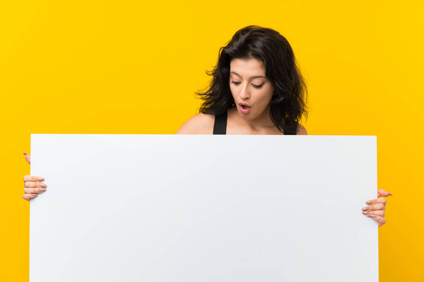 Jonge vrouw over geïsoleerde gele achtergrond met een lege witte plakkaat voor invoegen een concept - Foto, afbeelding