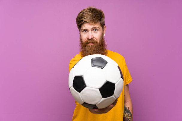 rothaariger Mann mit langem Bart, der einen Fußball über eine isolierte lila Wand hält - Foto, Bild