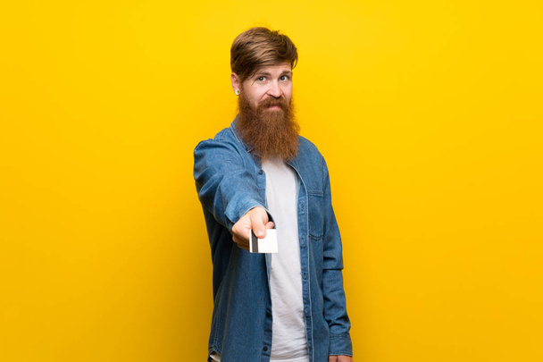 Pelirrojo con barba larga sobre pared amarilla aislada sosteniendo una tarjeta de crédito
 - Foto, imagen