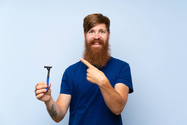 rothaarige Mann rasiert seinen Bart über isolierten blauen Hintergrund zeigt auf die Seite, um ein Produkt zu präsentieren - Foto, Bild