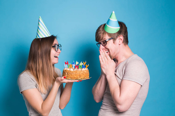 Vrolijke jonge paar charmante kerel en schattig meisje in papier hoeden maken dwaze gezicht en vasthouden in hun handen een verjaardagstaart staande op een blauwe achtergrond. - Foto, afbeelding