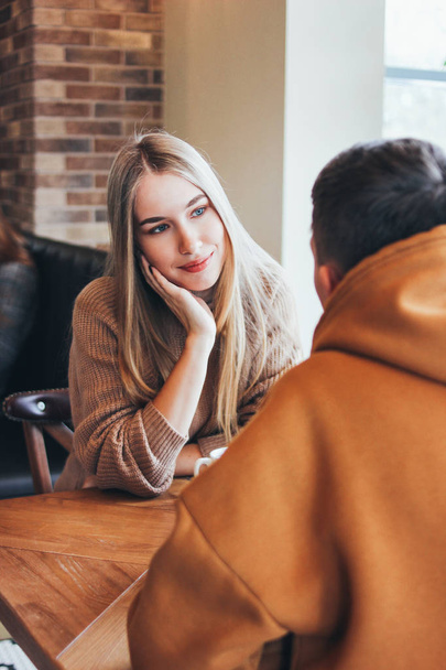 Beautiful long hair blonde girl looking at boyfriend and smiling. Young couple dressed warm casual clothing sitting at cafe together - Photo, Image