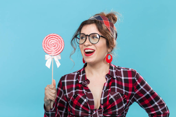 Közeli portréja egy bájos fiatal pin-up nő egy kockás inget és poharakat is nyalogatja a nagy nyalóka szemben a kék háttérben. Koncepció a szeretet édesség és desszert. Copyspace. - Fotó, kép