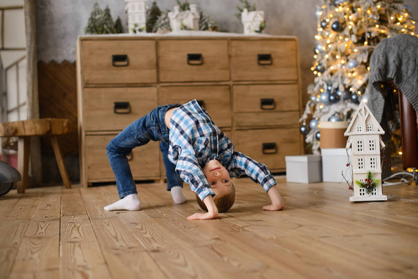boy stands in an acrobatic pose on  wooden floor, against background of Christmas tree, glowing with lights and bokeh - Photo, Image
