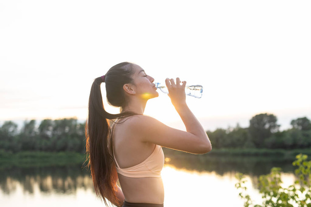 Όμορφη αθλήτρια γυμναστικής γυναίκα πόσιμο νερό μετά την προπόνηση άσκηση στο ηλιοβασίλεμα το βράδυ του καλοκαιριού στην παραλία εξωτερική πορτρέτο - Φωτογραφία, εικόνα