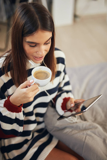 Hermosa morena caucásica vestida con suéter sentada en la cama en el dormitorio, usando tableta y tomando café por la mañana
. - Foto, Imagen