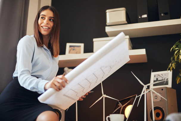Attractive businesswoman in shirt and skirt sitting on the desk in office and and plans. Sustainable development concept. - Фото, зображення