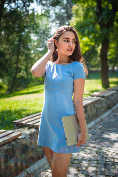 Jonge brunette student in een blauwe jurk neemt een wandeling in een park op een warme zonnige dag. Leven op de campus. - Foto, afbeelding