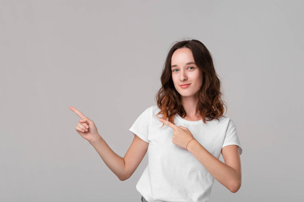 Pretty brunette woman in a white tee pointing at the empty space in a left side of a frame standing isolated over grey background. Copy space of advertising. - Foto, imagen