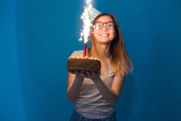Cheerful young blurred girl student in glasses holding a congratulatory cake with a candle standing on a blue background. Birthday concept. - Photo, Image