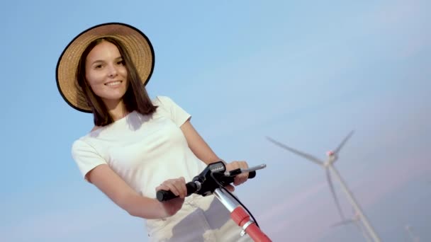 Ritratto di ragazza in un elegante cappello di paglia con scooter elettrico sullo sfondo di parchi eolici
. - Filmati, video