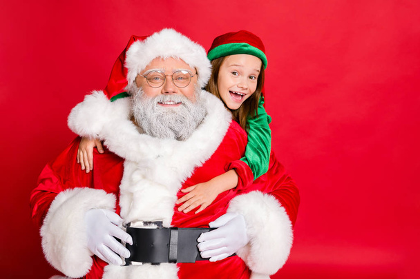 Close up photo of charming santa claus in eyeglasses eyewear and his little elf helper in green hat headwear hugging piggyback isolated over red background - Fotó, kép