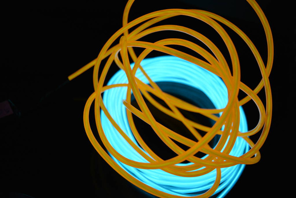 eine helle Spule aus türkisfarbenem, himmelblauem Leuchtdraht mit chaotischen Drähten eines orangefarbenen Lichtleiters, der sich auf einer schwarz glänzenden Oberfläche befindet. Lichtleinwand, Hintergrundbeleuchtung mit Drähten und Kunsthintergrund. - Foto, Bild