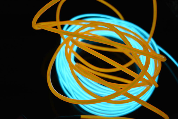 Яркая катушка бирюзовой, небесно-голубой светящейся проволоки с хаотичными проводами оранжевого света, расположенной на черной глянцевой поверхности. Легкий холст, подсветка с проводами и художественным фоном
. - Фото, изображение