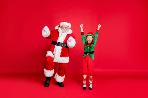 Portrait of cute santa claus elf in green costumes with hat headwear eyewear eyeglasses raising fists screaming yes have newyear party isolated over red background - 写真・画像