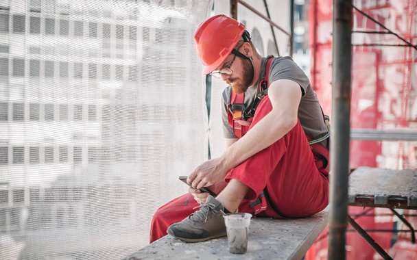 Rakennustyömaan työntekijä rakennustelineissä matkapuhelimen avulla
 - Valokuva, kuva