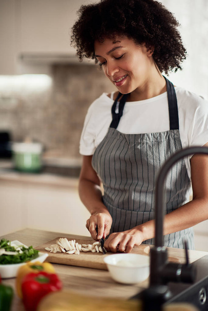 Gorgeous mixed race housewife in apron standing in kitchen and chopping mushrooms. On kitchen counter are all sorts of vegetables. - Foto, Imagen