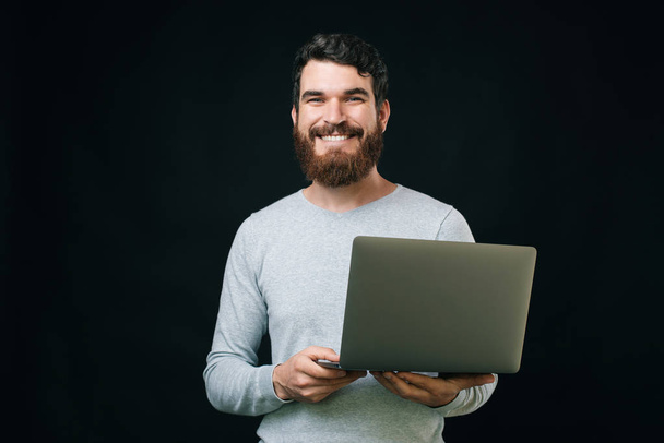 Foto van de jonge lachende man houdt laptop en kijken naar de camera over donkere achtergrond - Foto, afbeelding