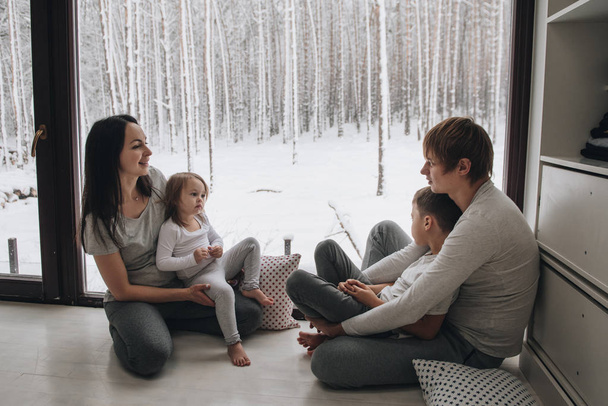 Семья сидит у окна и смотрит на зимний лес. Хороший новогодний настрой. Утро в пижаме
. - Фото, изображение