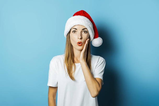 Bella giovane donna con una faccia sorpresa in un cappello di Babbo Natale su uno sfondo blu. Idea concettuale per Capodanno e Natale
 - Foto, immagini