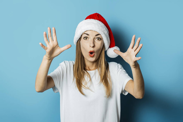 Hermosa mujer joven con una cara sorprendida en un sombrero de Santa Claus sobre un fondo azul. Concepto de idea para el año nuevo y la Navidad
 - Foto, imagen
