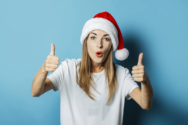 Beautiful Young woman in Santa Claus hat makes hand gestures on a blue background. Concept idea for New Year and Christmas, Christmas presents - Photo, Image