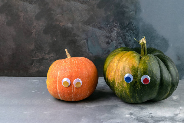 Green and orange pumpkins with eyes on grey cement background. Autumn creative concept with copy space - Фото, изображение